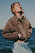 Person wearing a brown jacket and light blue pants standing by the ocean.