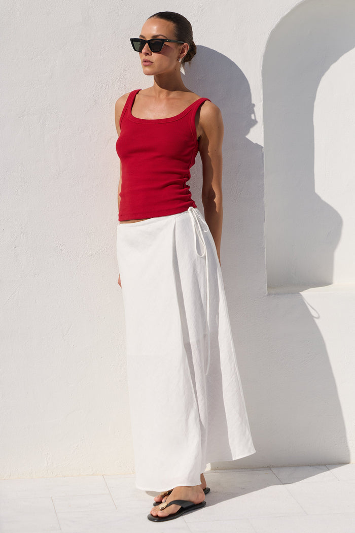 A woman wearing a red ribbed tank top and a long white skirt, standing against a white wall with strong sunlight and shadows.