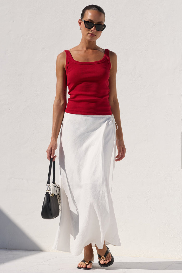 A woman stands outdoors against a bright white wall wearing a sleeveless red top and a long white skirt, holding a black handbag.