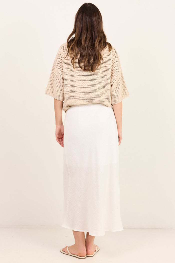 A woman viewed from behind wearing a beige textured short-sleeve top and a long white skirt, standing against a plain off-white studio background.