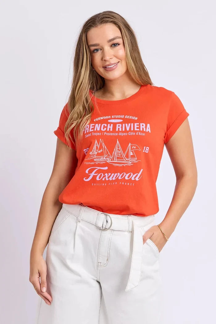A woman modeling a bright orange short-sleeve graphic t-shirt with white text and sailboat illustrations reading 'French Riviera' and 'Foxwood', paired with white pants, against a light gray studio background.