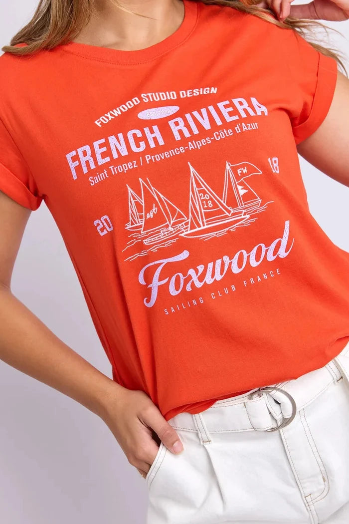 A woman wearing a bright orange short-sleeve graphic T-shirt with white printed text reading 'French Riviera' and sailboat artwork, paired with white pants against a light gray studio background.