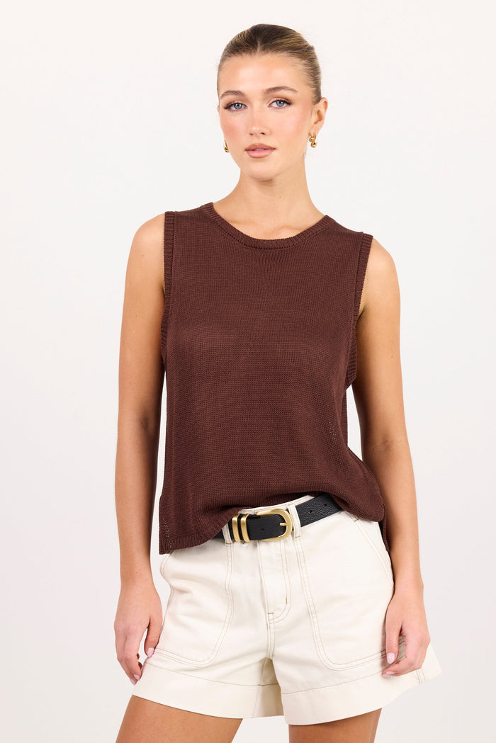 A woman modeling a sleeveless dark brown knit top paired with off-white shorts and a black belt with a gold buckle, set against a plain light gray studio background.