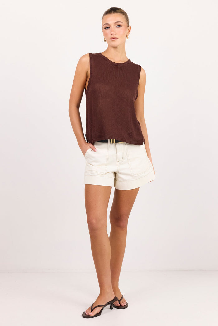 A woman modeling a sleeveless dark brown knit top paired with off-white shorts and black sandals, standing against a plain white studio background.