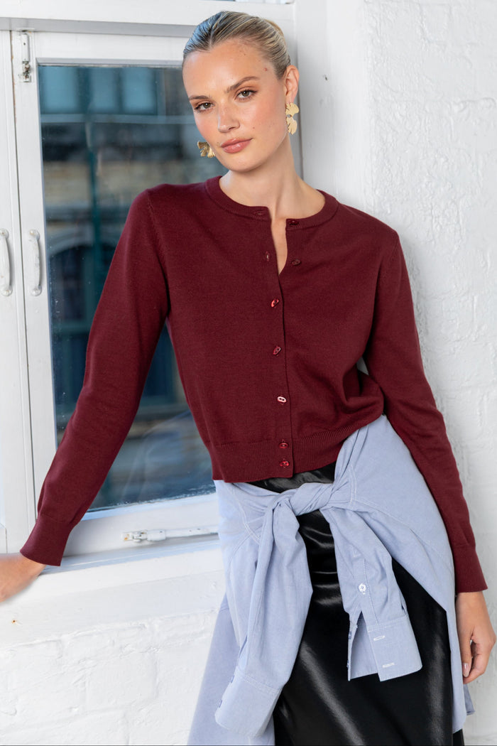 Woman in a burgundy shirt leaning against a window with a white wall background