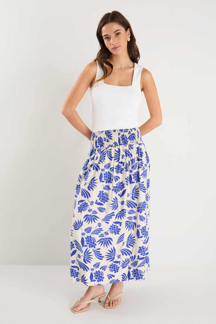 A model wears a long white skirt with a bold blue floral print, elastic waistband, and relaxed fit, paired with a white tank top and neutral sandals.