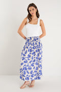 A model wears a long white skirt with a bold blue floral print, elastic waistband, and relaxed fit, paired with a white tank top and neutral sandals.