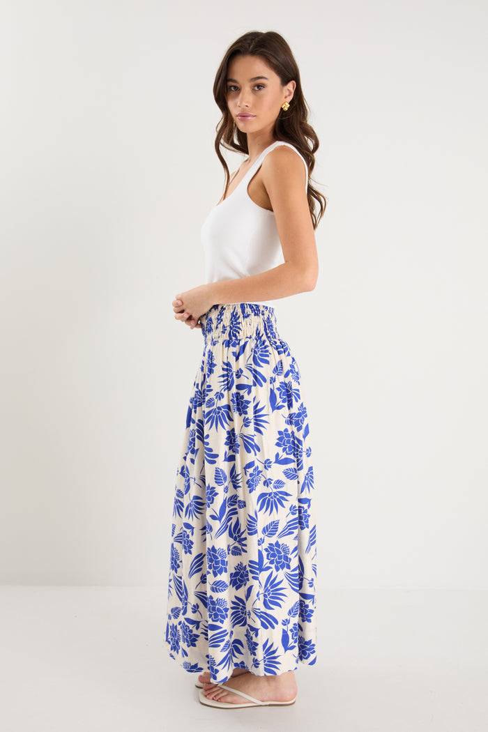 A model wears a long white skirt with a bold blue floral print, elastic waistband, and relaxed fit, paired with a white tank top and neutral sandals.
