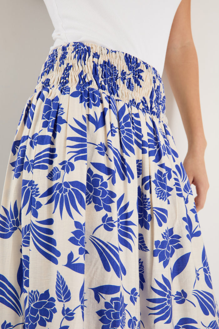 A model wears a long white skirt with a bold blue floral print, elastic waistband, and relaxed fit, paired with a white tank top and neutral sandals.