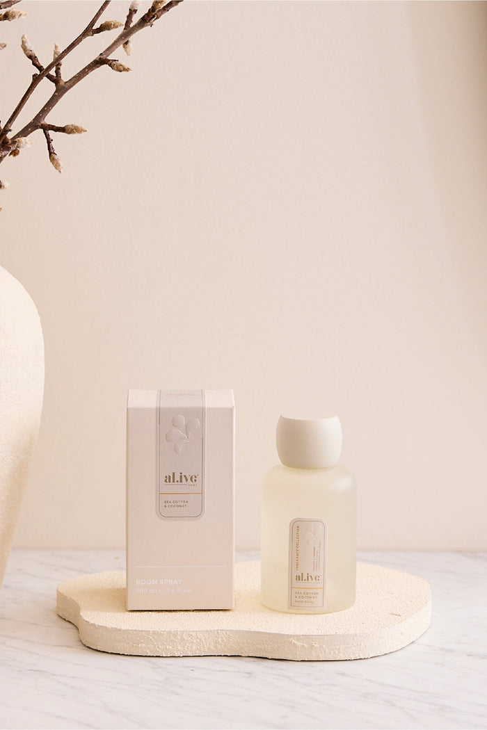 White candle and packaging on a wooden tray with a vase and branches in the background.