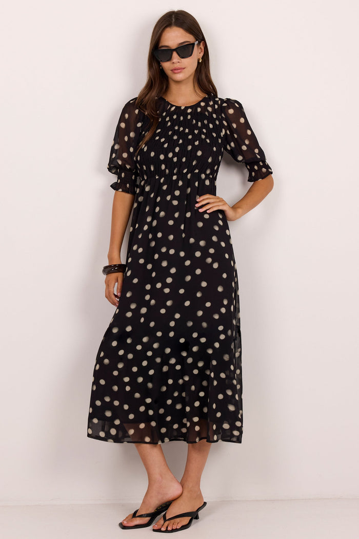 A woman modeling a black midi dress with a beige polka dot pattern, featuring short puff sleeves and a round neckline, set against a plain white studio background.
