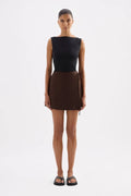 A model stands against a light gray studio background wearing a sleeveless black top and a brown wrap-style mini skirt with side ties, paired with black sandals.