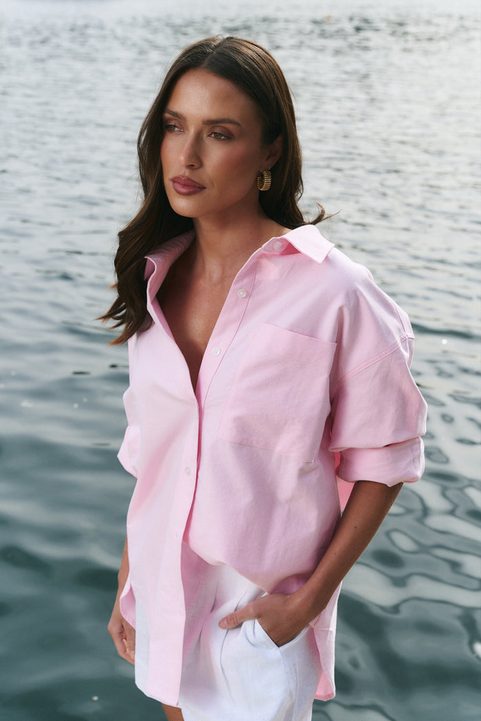 Woman in a pink shirt and white shorts standing by a body of water with boats in the background.