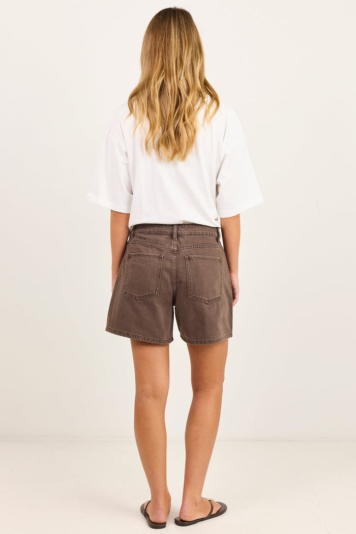 A woman viewed from behind wearing brown denim shorts, a white t-shirt, and black sandals, standing against a plain off-white studio background.