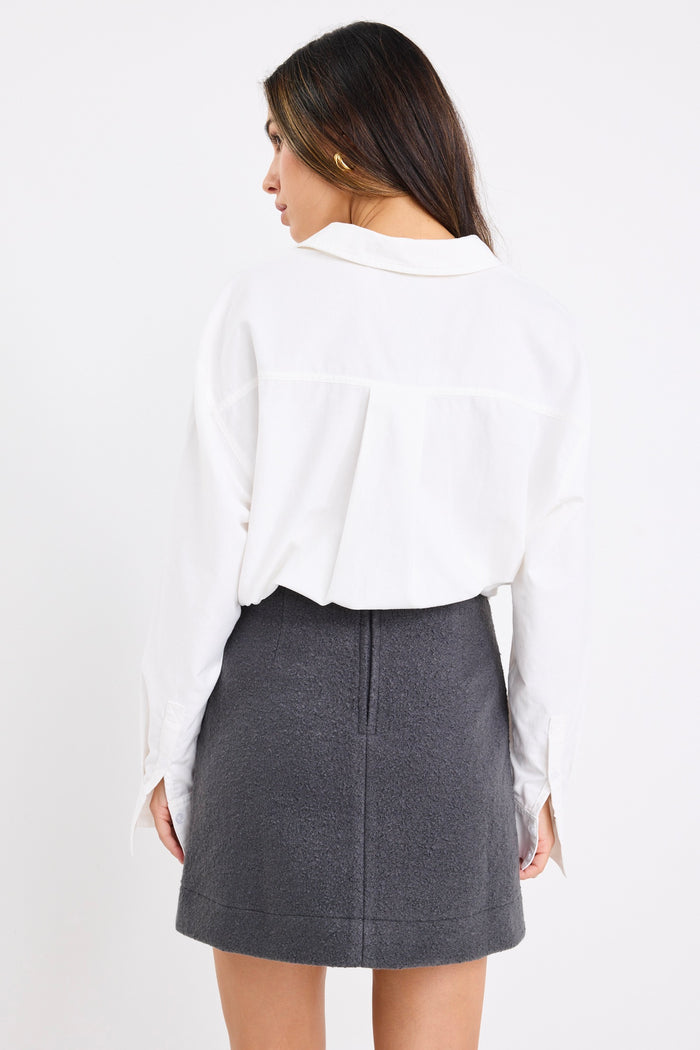 Woman wearing a dark charcoal grey mini skirt with a white oversized shirt and black sneakers, showing a clean, minimal outfit with street-style flair.