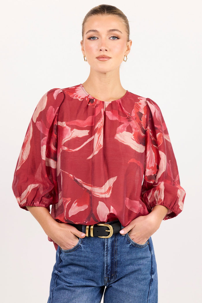 A woman modeling a red and pink abstract print blouse with voluminous puff sleeves, paired with blue jeans and a black belt, against a plain white studio background.