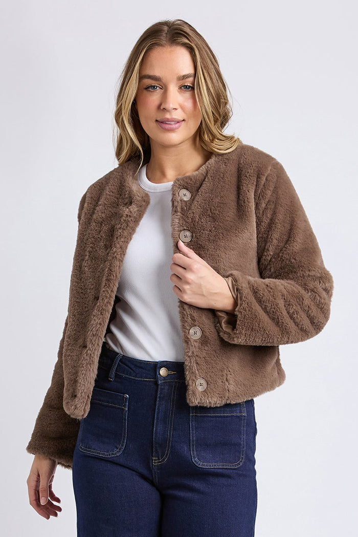 Woman wearing a brown jacket, white shirt, blue jeans, and white sneakers on a light gray background