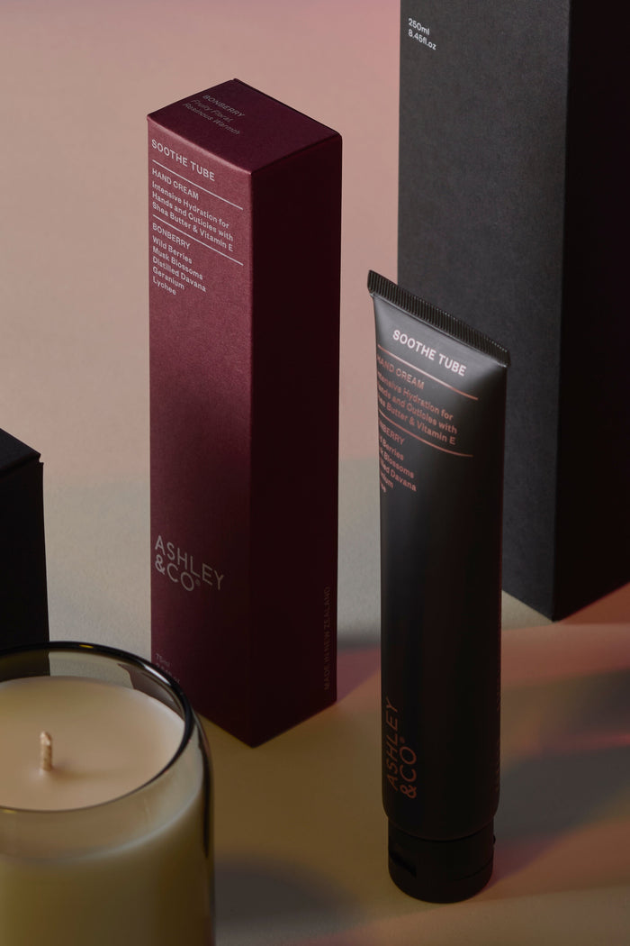 Candle, box, and tube of Ashley & Co. product on a neutral background