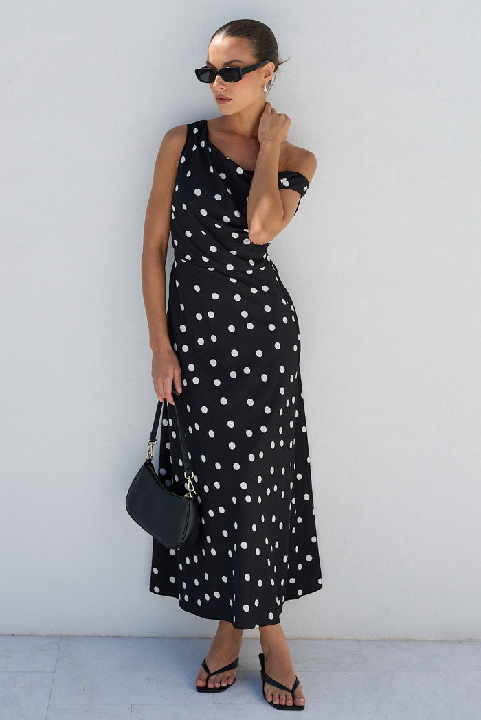 A woman modeling a black sleeveless maxi dress with white polka dots, standing against a minimalist white stucco wall.