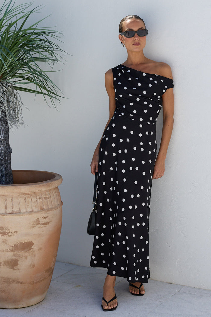 A woman wearing a black off-the-shoulder maxi dress with white polka dots, standing against a white stucco wall next to a large potted palm.