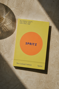 A book titled "SPRITZ" by Spritz, featuring a minimalist yellow cover with a large orange circle and blue text, photographed on a textured beige stone surface with dramatic shadows.
