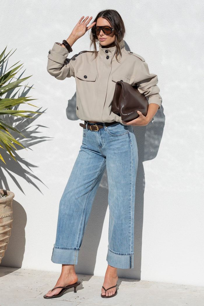 A woman stands outdoors against a white wall wearing a light beige utility jacket, light blue wide-leg jeans, and black sandals, holding a brown leather clutch. A large potted yucca plant is to her left.