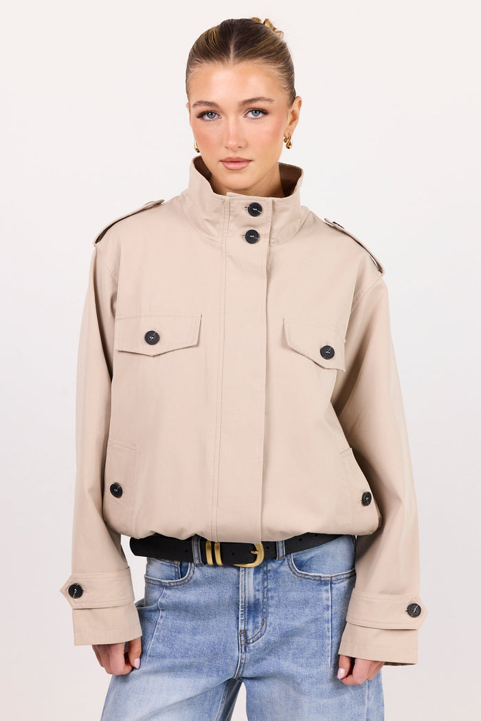 A woman modeling a cropped beige military-style jacket with a high stand collar, flap chest pockets, and black buttons, paired with light blue jeans and a black belt, against a plain off-white studio background.