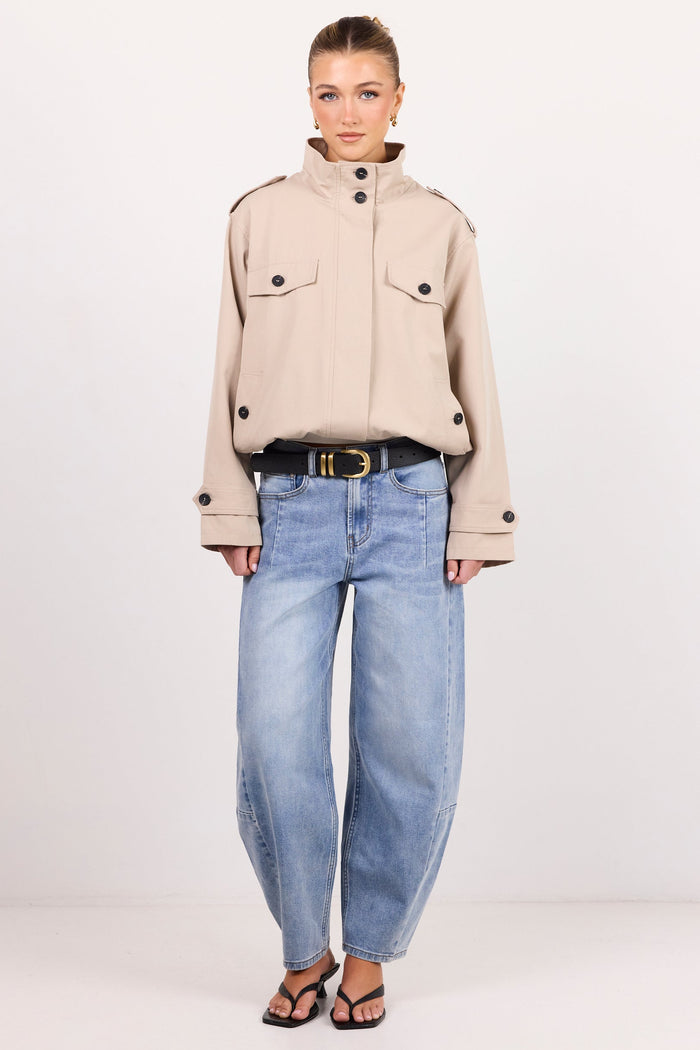 A model stands against a plain white studio background wearing a beige cropped military-style jacket with black buttons, light-wash blue wide-leg jeans, a black belt with a gold buckle, and black sandals.