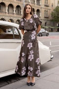 Woman in a floral dress standing next to a vintage car on a city street.