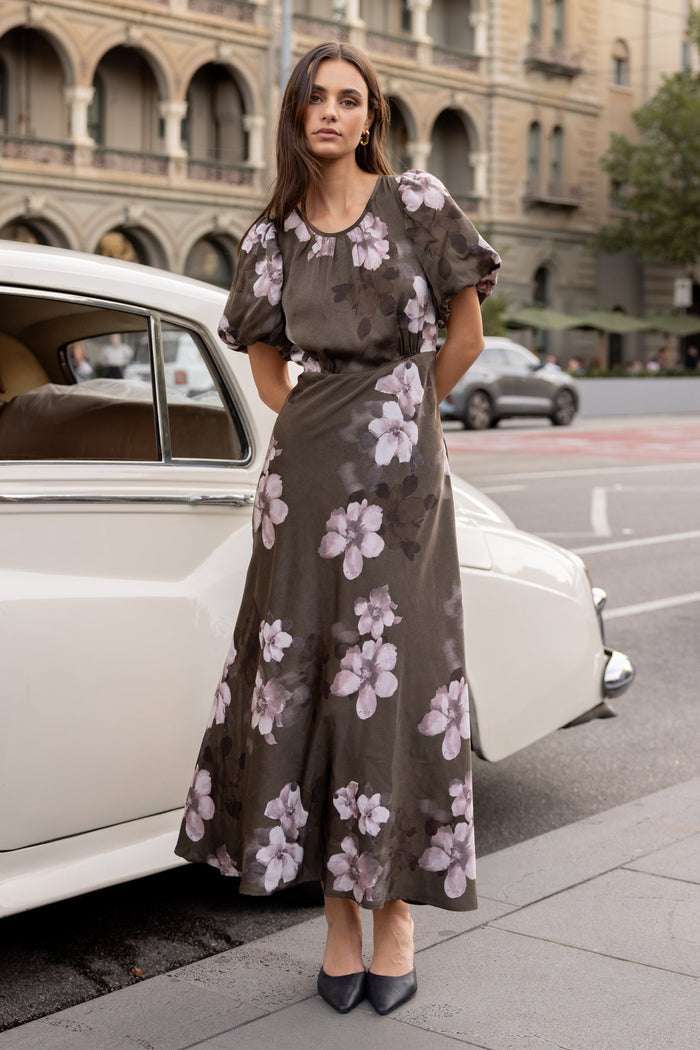 Woman in a floral dress standing next to a vintage car on a city street.