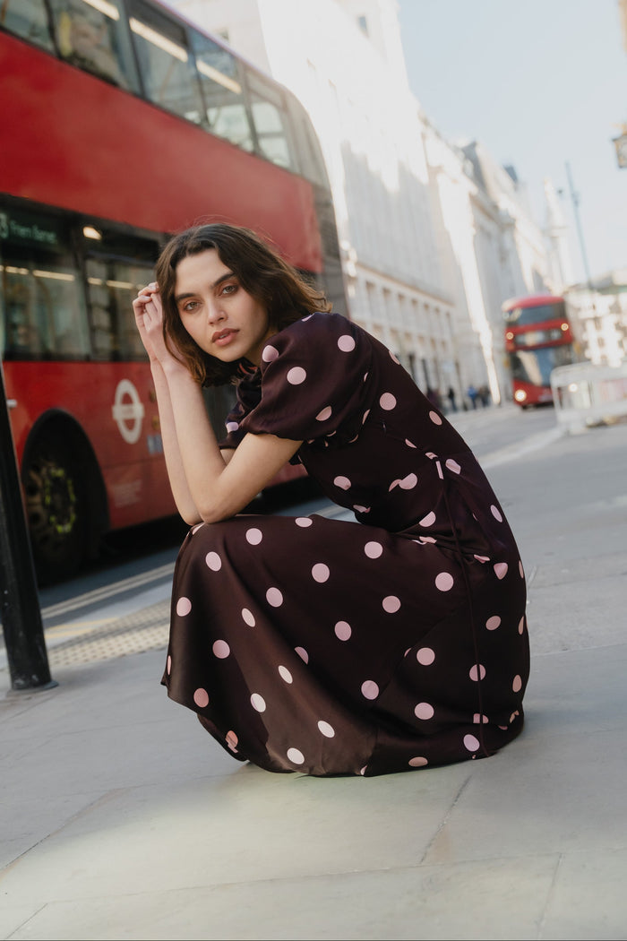 Woman in a polka dot dress sitting on a city street with red buses in the background