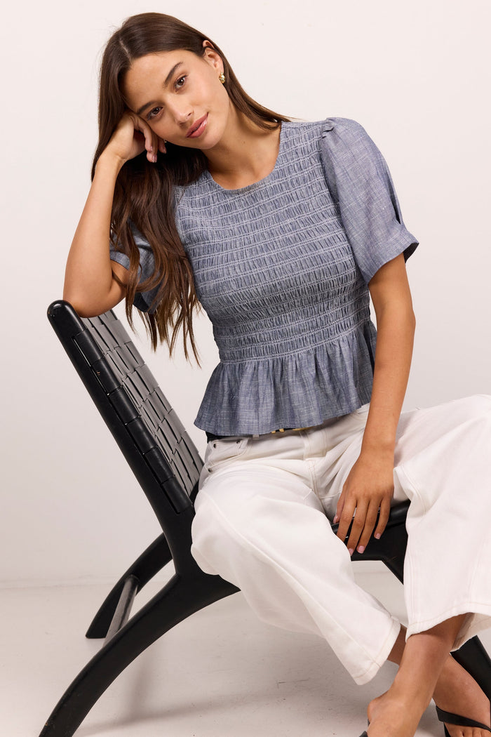 A woman models a blue-gray smocked short-sleeve peplum top paired with white cropped pants and black sandals, seated on a black modern chair against a plain off-white studio background.