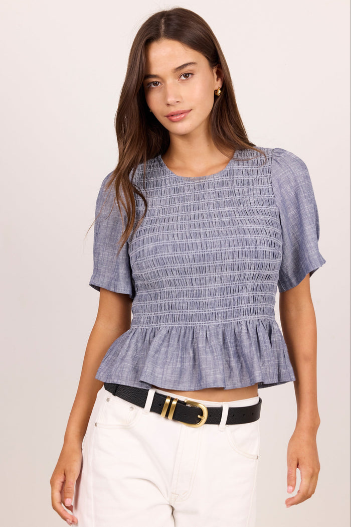 A woman modeling a short-sleeve smocked blouse in a dusty periwinkle blue color, paired with white pants and a black belt, against a plain light beige background.
col_pattern