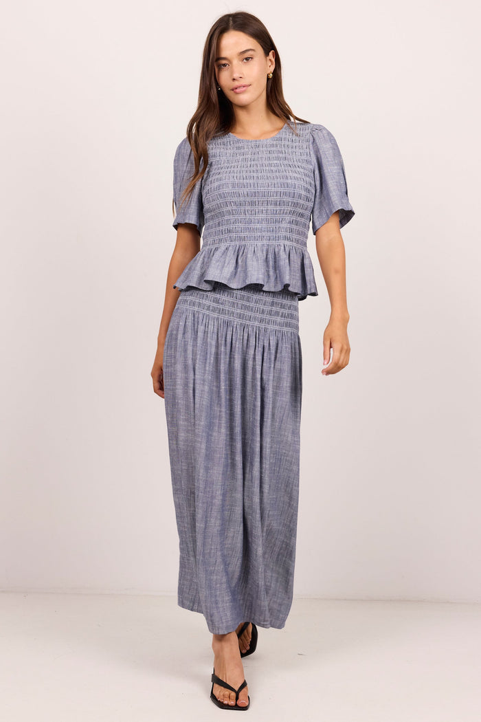 A woman modeling a matching two-piece set consisting of a smocked short-sleeve top with a ruffle hem and matching wide-leg pants in a textured slate blue fabric, set against a plain light background.