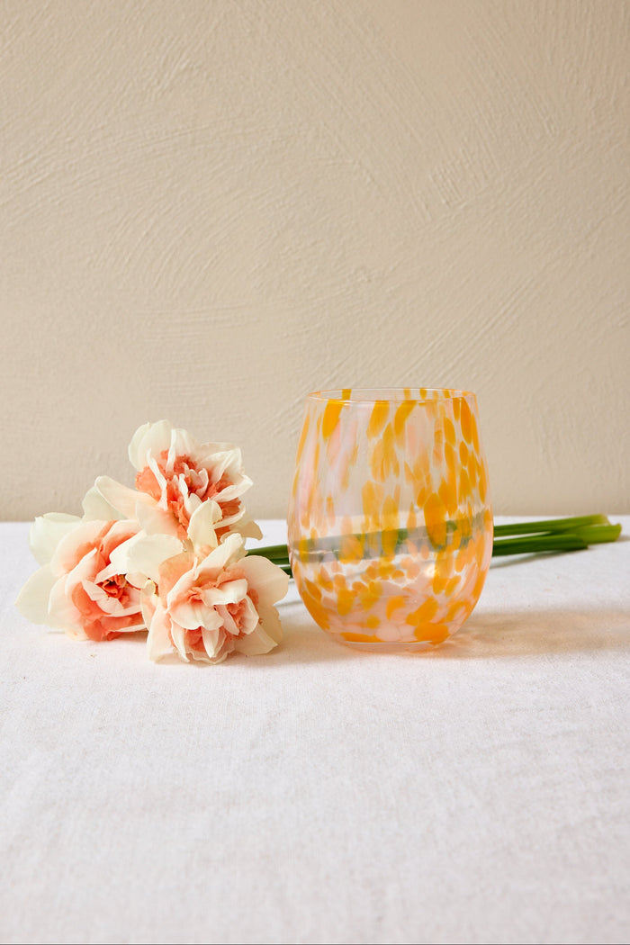 Glass with yellow and orange pattern next to pink flowers on a white surface