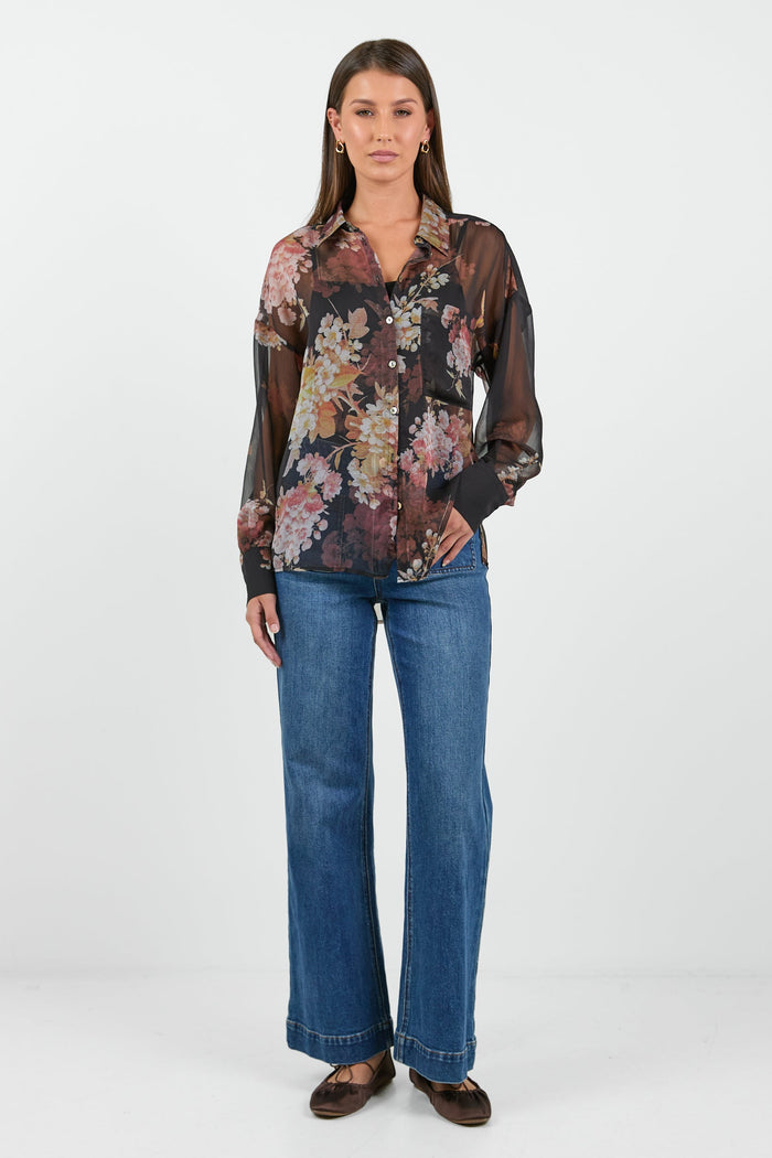 A woman wearing a black sheer blouse with a floral bouquet print in shades of pink, orange, and cream.