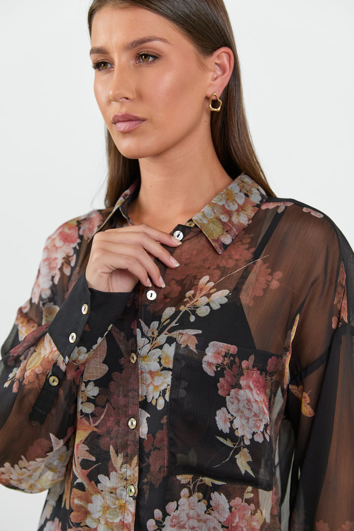 A woman wearing a black sheer blouse with a floral bouquet print in shades of pink, orange, and cream.