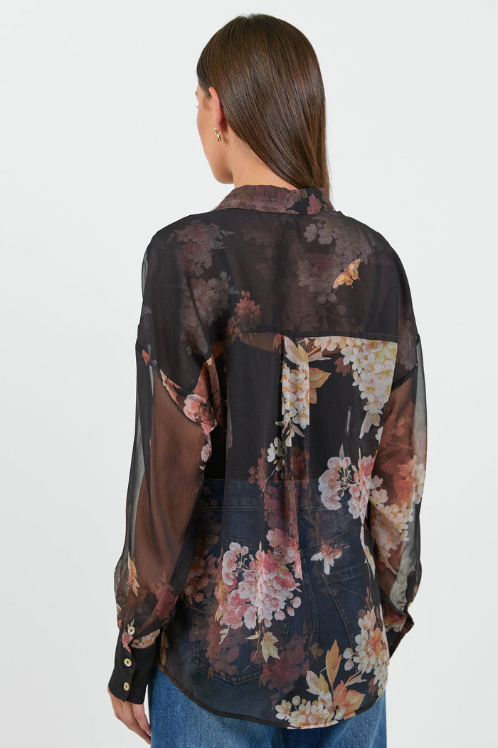 A woman wearing a black sheer blouse with a floral bouquet print in shades of pink, orange, and cream.
