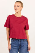 A woman modeling a short-sleeve crewneck t-shirt in a deep red color, paired with blue jeans, against a plain off-white studio background.