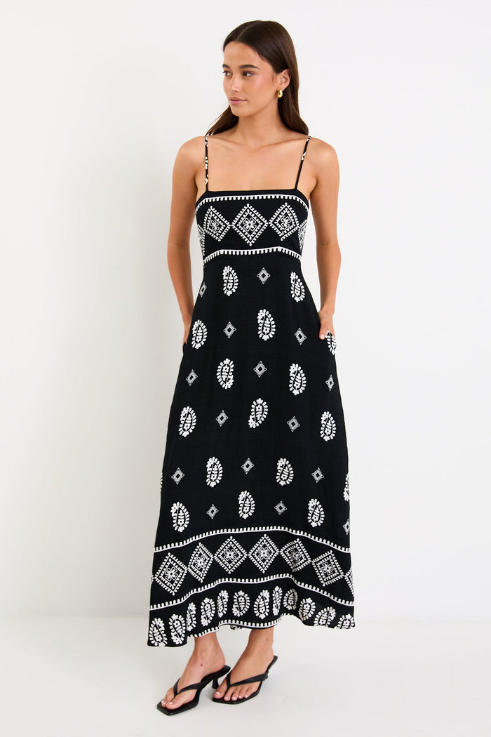 Woman wearing a black maxi dress with fine straps, white paisley and border print, and shirred back detail.