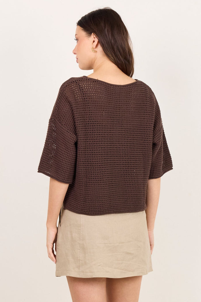 A woman is shown from the back wearing a dark brown textured short-sleeve top and a beige mini skirt, standing against a plain off-white studio background.