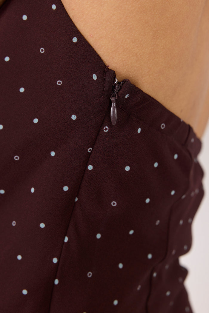 Close-up of a dark brown or very deep burgundy fabric with a small white polka dot pattern, showing a zipper detail and part of a model's skin.