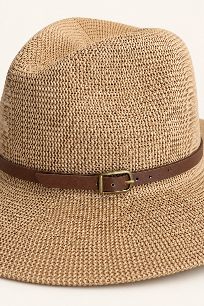 A close-up of a woven straw fedora hat in a warm tan color, featuring a brown leather band with a brass buckle.