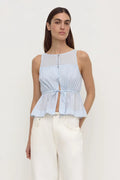 A woman modeling a light blue sleeveless top with a sheer yoke, button front, and gathered peplum hem, paired with white pants, against a plain light gray studio background.