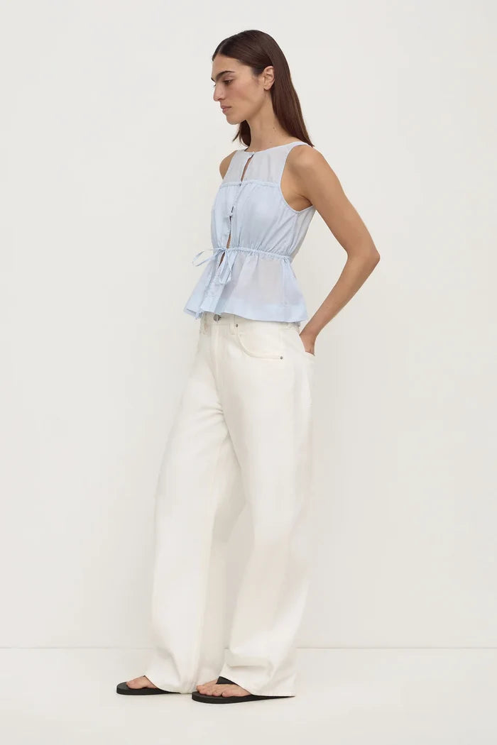A woman modeling a light blue sleeveless peplum top with a tie waist, paired with white wide-leg trousers and black sandals, standing against a plain white studio background.