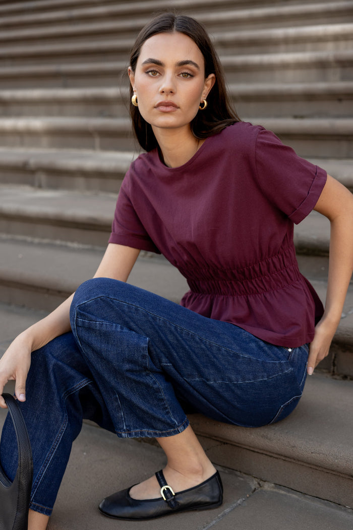 Woman sitting on steps wearing a purple top and blue jeans.