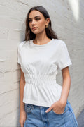 Woman wearing a white top and blue jeans leaning against a light gray brick wall.