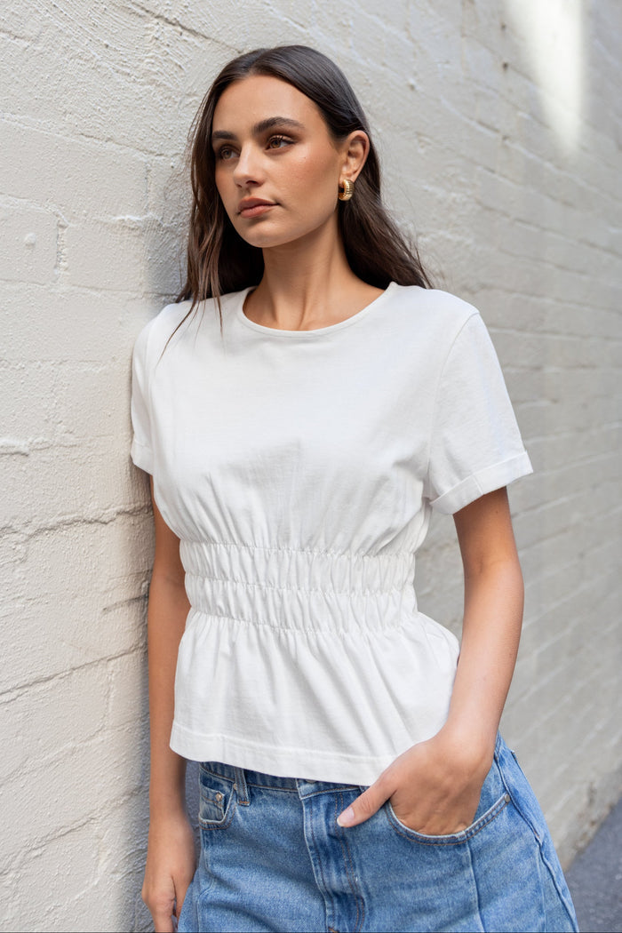 Woman wearing a white top and blue jeans leaning against a light gray brick wall.