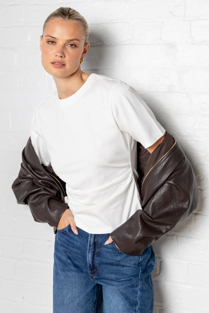 Woman wearing a white t-shirt with side ruching, leather bomber jacket and blue jeans against a plain background
