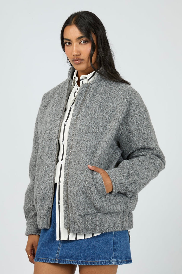 Model wears a grey marle bomber jacket with a textured wool-blend finish. The jacket features a high collar, front zip closure, and relaxed fit with side pockets. Layered over a striped button-down shirt and styled with a denim mini skirt for a casual yet elevated outfit.